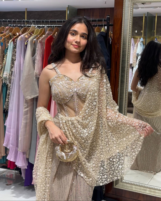 A woman modeling a heavily embellished beige and gold traditional outfit with a matching sheer embroidered shawl, standing in a clothing boutique with dress racks and a mirror in the background.