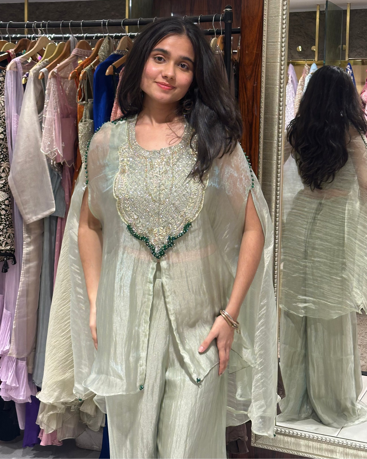 A woman modeling a light sage green embroidered traditional outfit with a sheer draped jacket, standing in a clothing boutique with a mirror and racks of garments in the background.